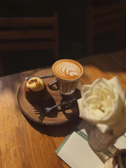 Coffee with latte art and a small cake on a sunlit wooden café table